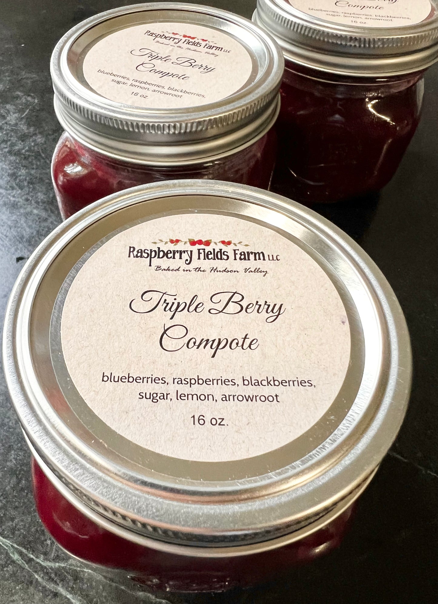 Three jars of Raspberry Fields Farm Triple Berry Compote on a dark surface.