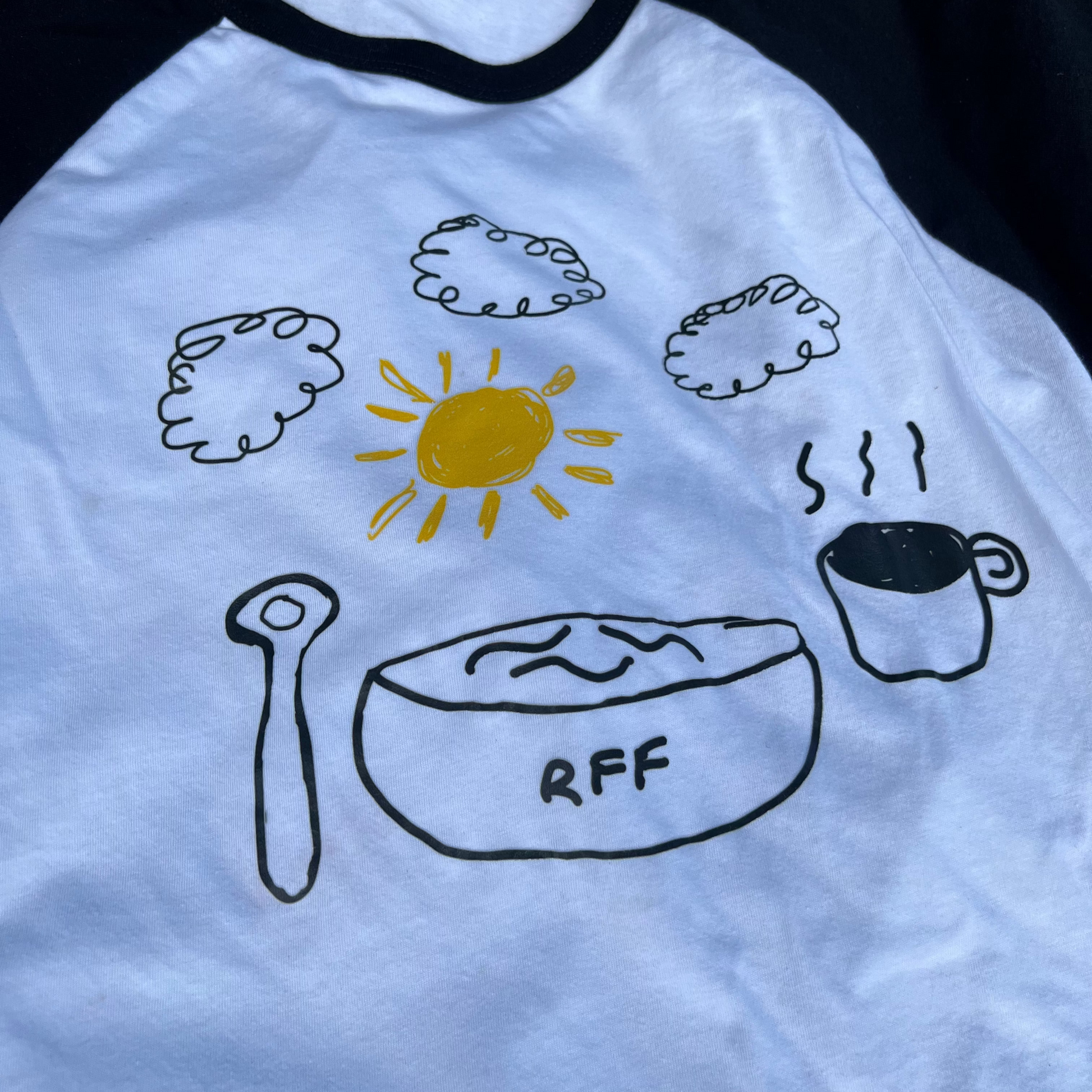 White t-shirt with black sleeves featuring hand-drawn illustrations of a sun, clouds, a bowl, and a cup on a grass background.