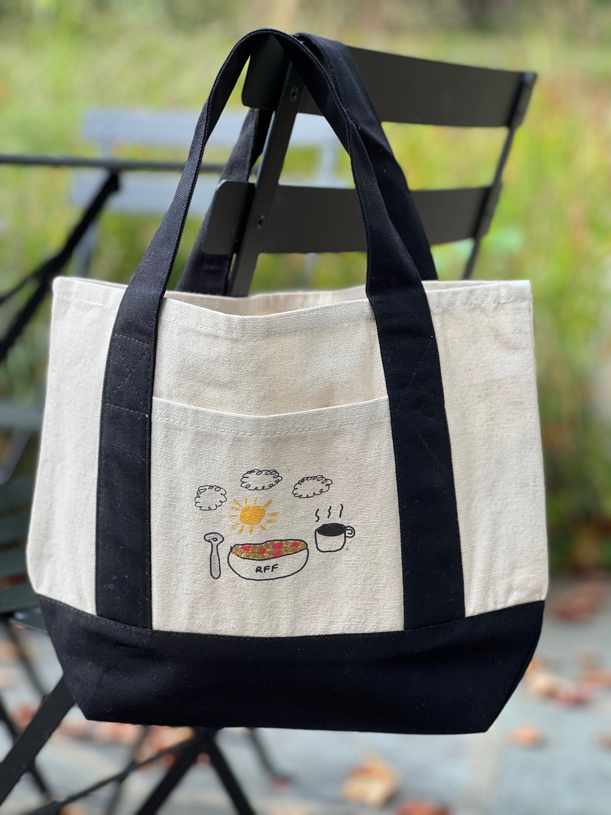 Tote bag with black handles and white front pocket featuring food illustrations, hanging on a chair outdoors.