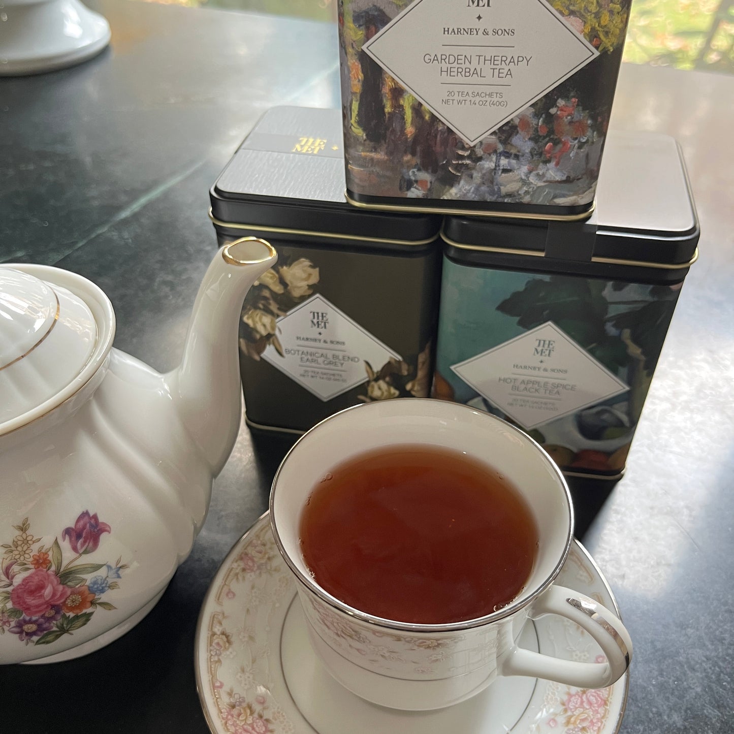 Tea cup with saucer and teapot on a reflective surface next to a tea box.
